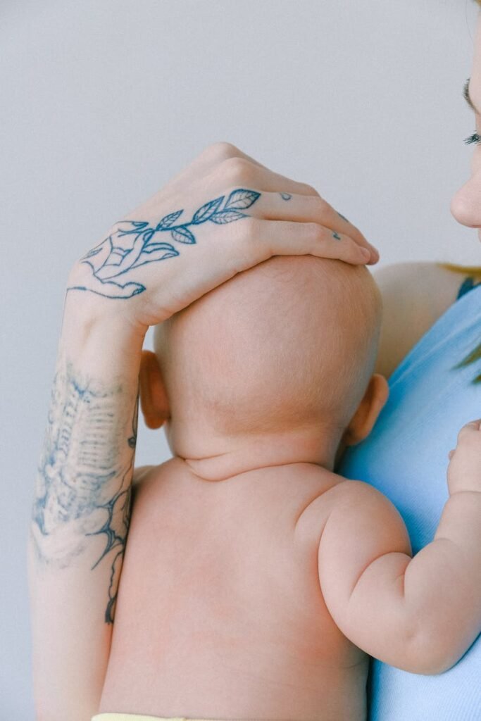crop mother with tattoo patting head of newborn while hugging toddler in room